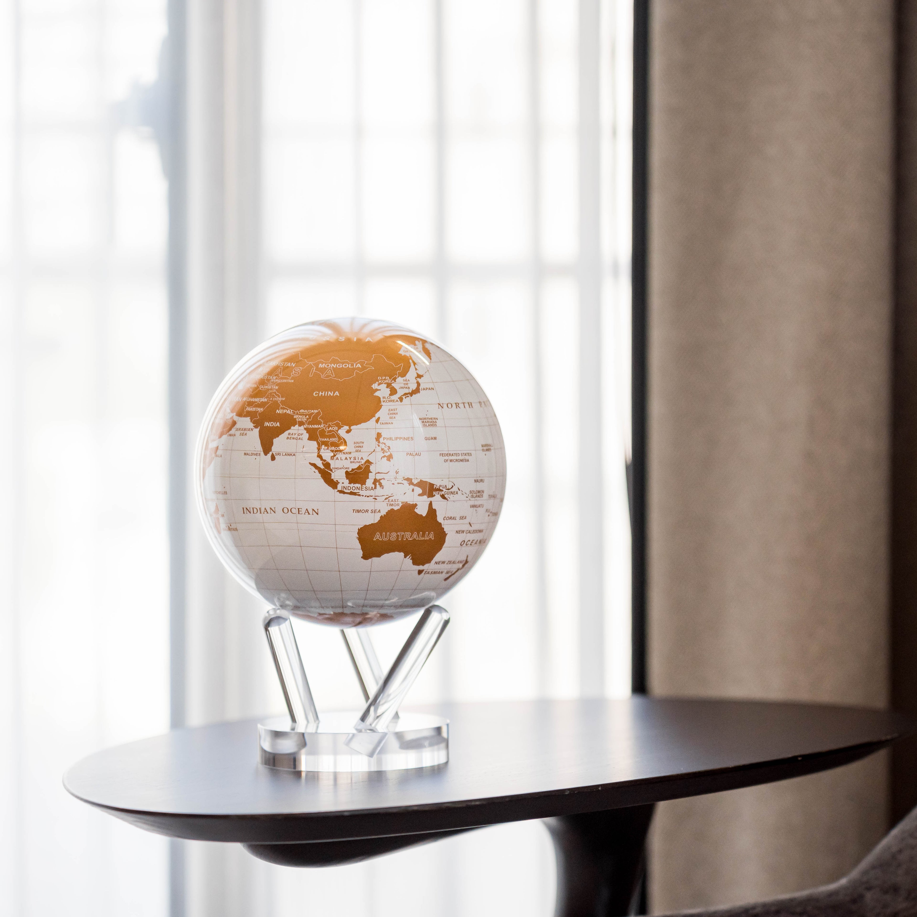 Pearl White & Gold MOVA Globe with acrylic base displayed by a sunlit window in a modern living room