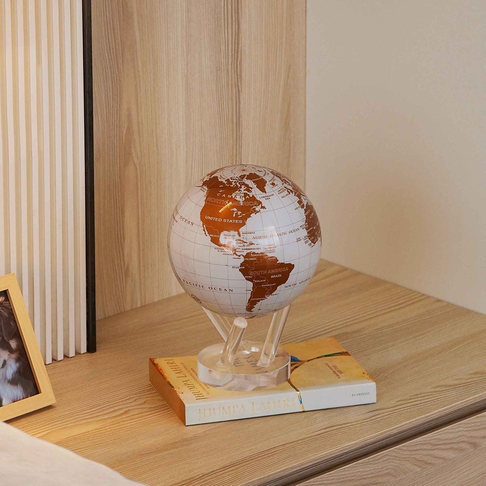 Pearl White & Gold MOVA Globe displayed on books in a modern wood shelving unit
