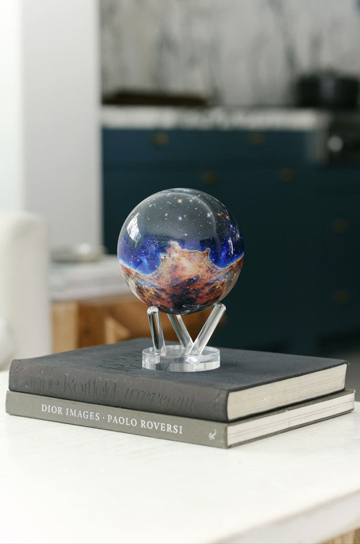 A stunning MOVA Globe featuring the James Webb Telescope’s vivid Carina Nebula image, displayed on a sleek clear stand atop two  books. 