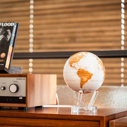 Pearl White &amp; Gold MOVA Globe on a wooden cabinet beside audio equipment and window blinds